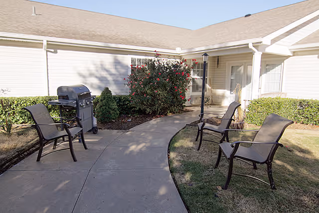 Outdoor patio area at Brookdale Owasso with a concrete walkway, four metal chairs with mesh seats and backs, a barbecue grill, and landscaping including bushes and a flowering shrub. The area is adjacent to a light-colored building with a shingled roof.