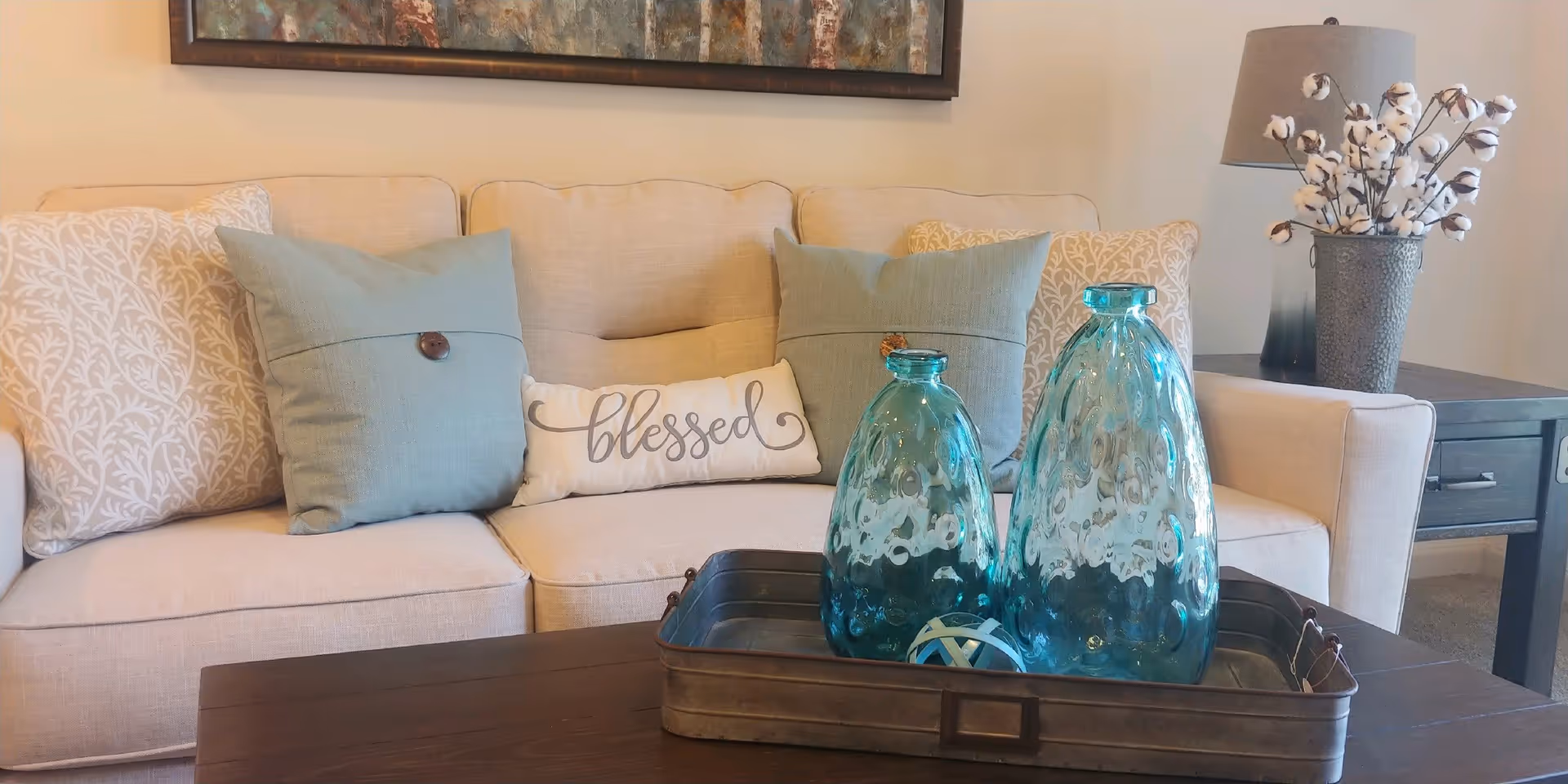 A beige sofa with decorative pillows, including two light blue pillows with buttons and a small white pillow with the word 'blessed' written on it. In front of the sofa is a dark wooden coffee table with a metal tray holding two blue glass vases and a decorative ball. To the right of the sofa is a side table with a lamp and a vase filled with cotton stems.