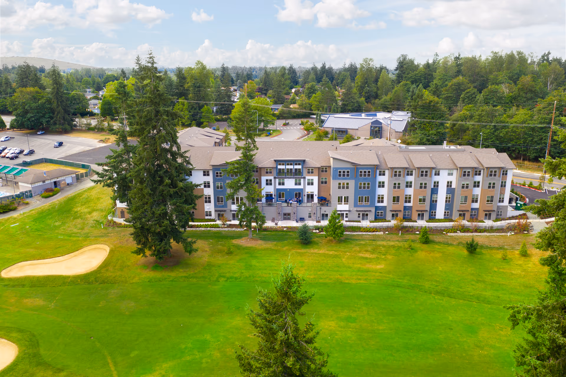 Aerial view of a multi-story senior living facility building surrounded by green lawns and trees, with a golf course sand trap visible in the foreground and a parking lot in the background under a partly cloudy sky.
