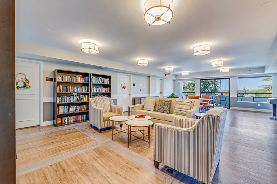 A bright and spacious senior living common area with comfortable seating including a sofa and two armchairs around a set of round coffee tables. There is a bookshelf filled with books against the wall, and large windows letting in natural light with views of trees outside. The room has modern ceiling lights and a wooden floor.