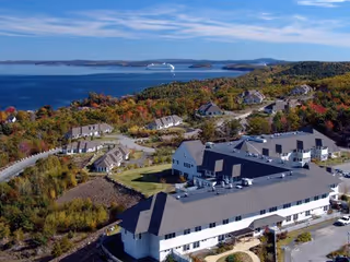 Aerial view of a retirement village situated near a large body of water with several buildings surrounded by trees and greenery. The landscape includes a mix of forested areas and open spaces, with a cruise ship visible on the water in the distance.