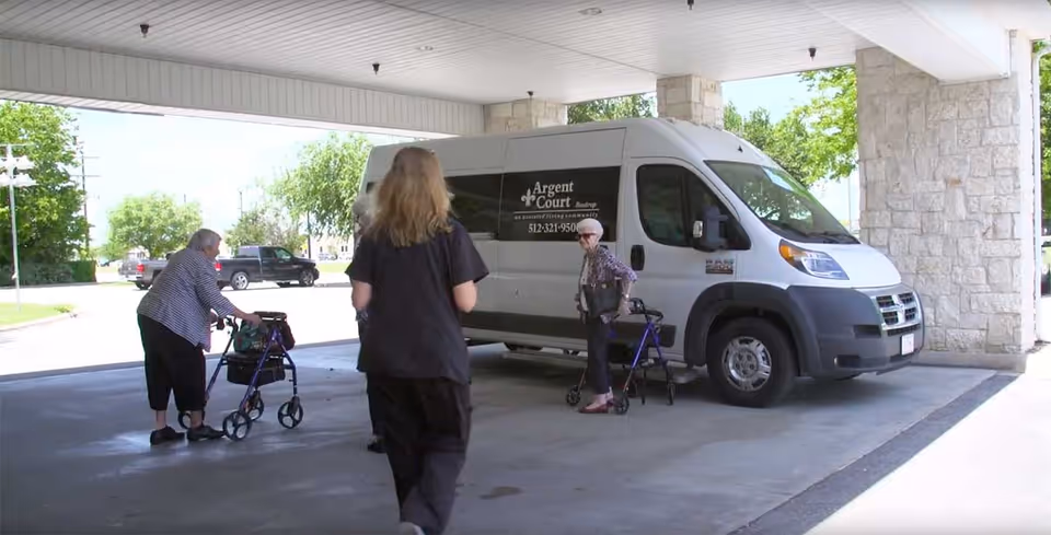Two elderly women with walkers and a caregiver standing under a covered driveway next to a white Argent Court Assisted Living van.