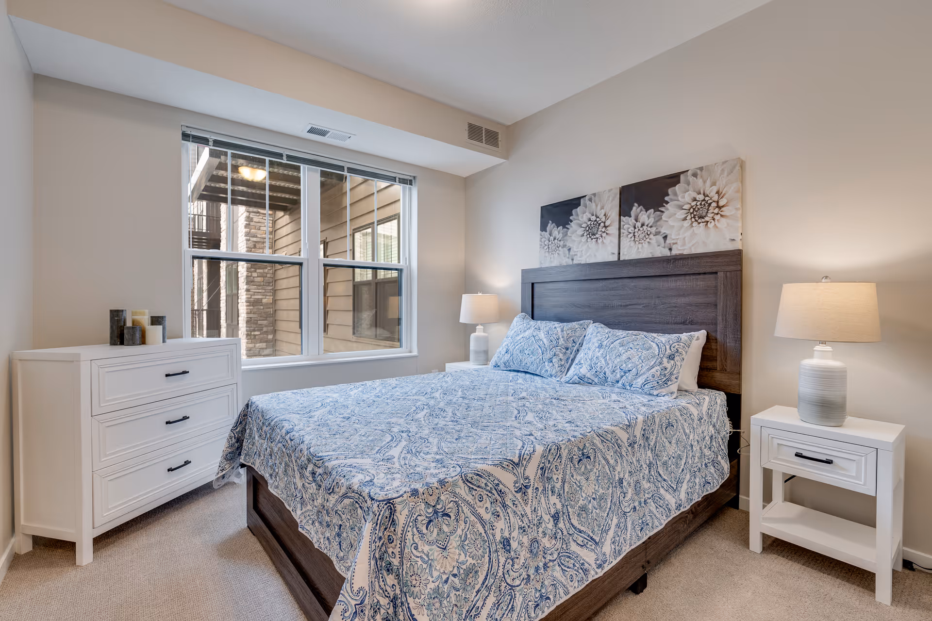 A bedroom with a large bed covered in a blue and white patterned bedspread and matching pillows. The bed has a dark wooden headboard with two floral artworks hanging above it. On either side of the bed are white nightstands with table lamps. There is a white dresser with decorative items on top near a window that shows an exterior view of a building with stone and wood siding.