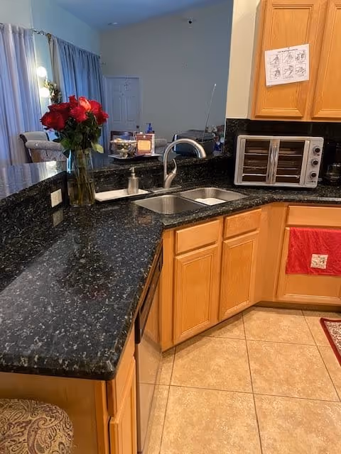 Interior view of a kitchen with black granite countertops, a double sink with a modern faucet, wooden cabinets, a small toaster oven, and a vase with red roses on the counter. The floor is tiled, and there is a red towel hanging on one of the cabinets.