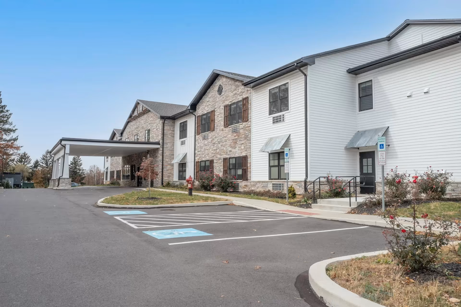 Exterior view of a senior living facility building with a covered entrance, stone and white siding facade, multiple windows, and a parking lot with designated handicapped parking spaces. There are small landscaped areas with bushes and flowers near the building.