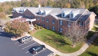 Aerial view of a red-brick, multi-story retirement community building with a curved driveway, parked cars, and landscaped lawn and trees.