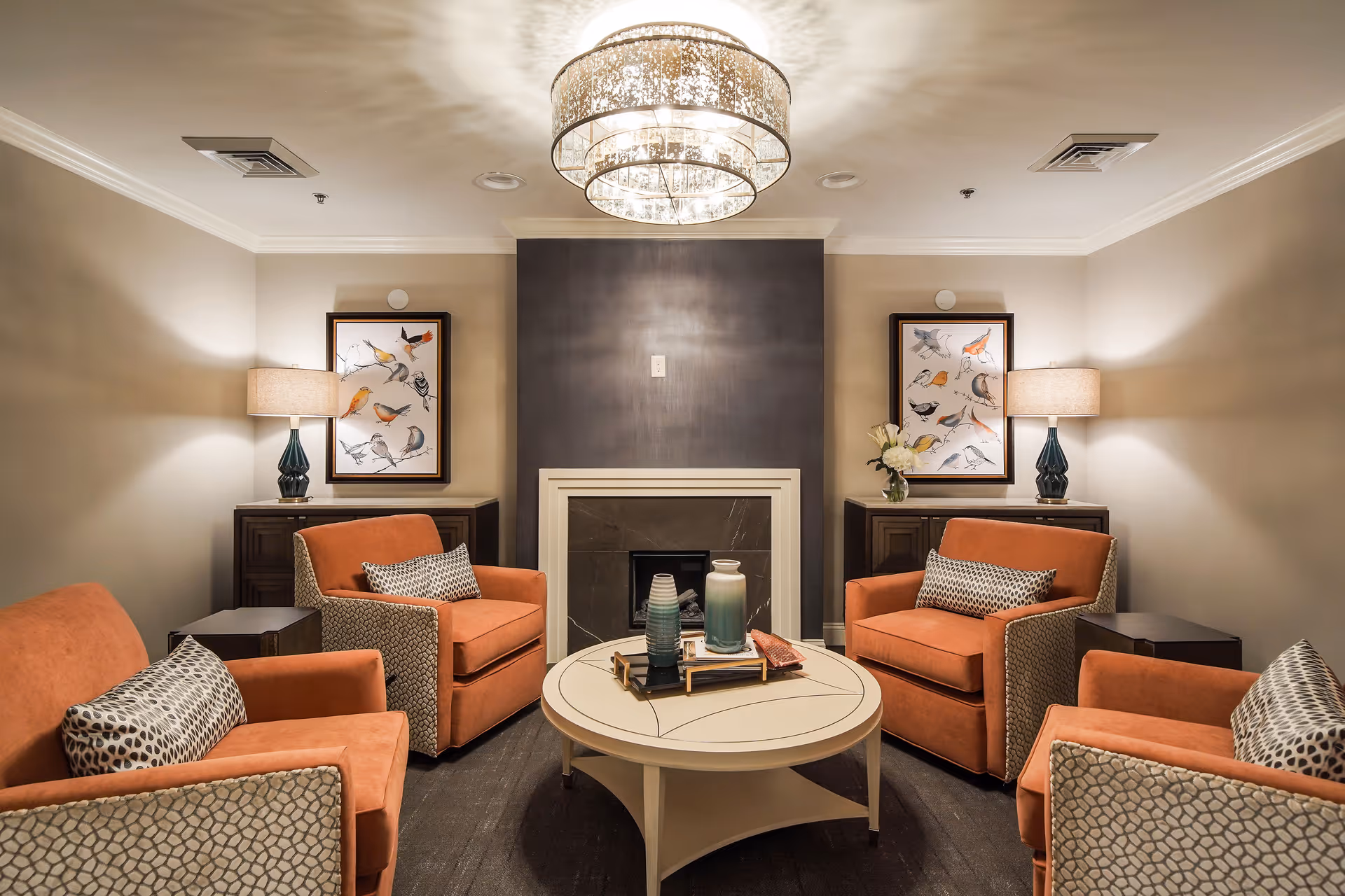 A cozy living room with four orange armchairs arranged around a round white coffee table. The table holds decorative vases and a tray. Behind the chairs, there is a dark fireplace with a white frame, flanked by two dark wooden cabinets each topped with a lamp and framed bird artwork. A modern chandelier hangs from the ceiling, casting light patterns on the ceiling.