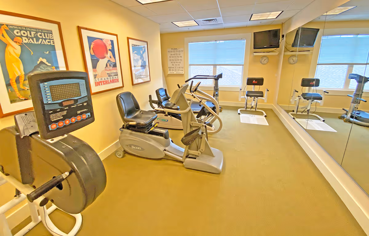 A small fitness room with exercise equipment including stationary bikes and a rowing machine. The room has beige walls, a large mirror on one side, two windows with blinds, and three framed vintage posters on the wall. There are also two small wall-mounted televisions and two clocks above the windows.