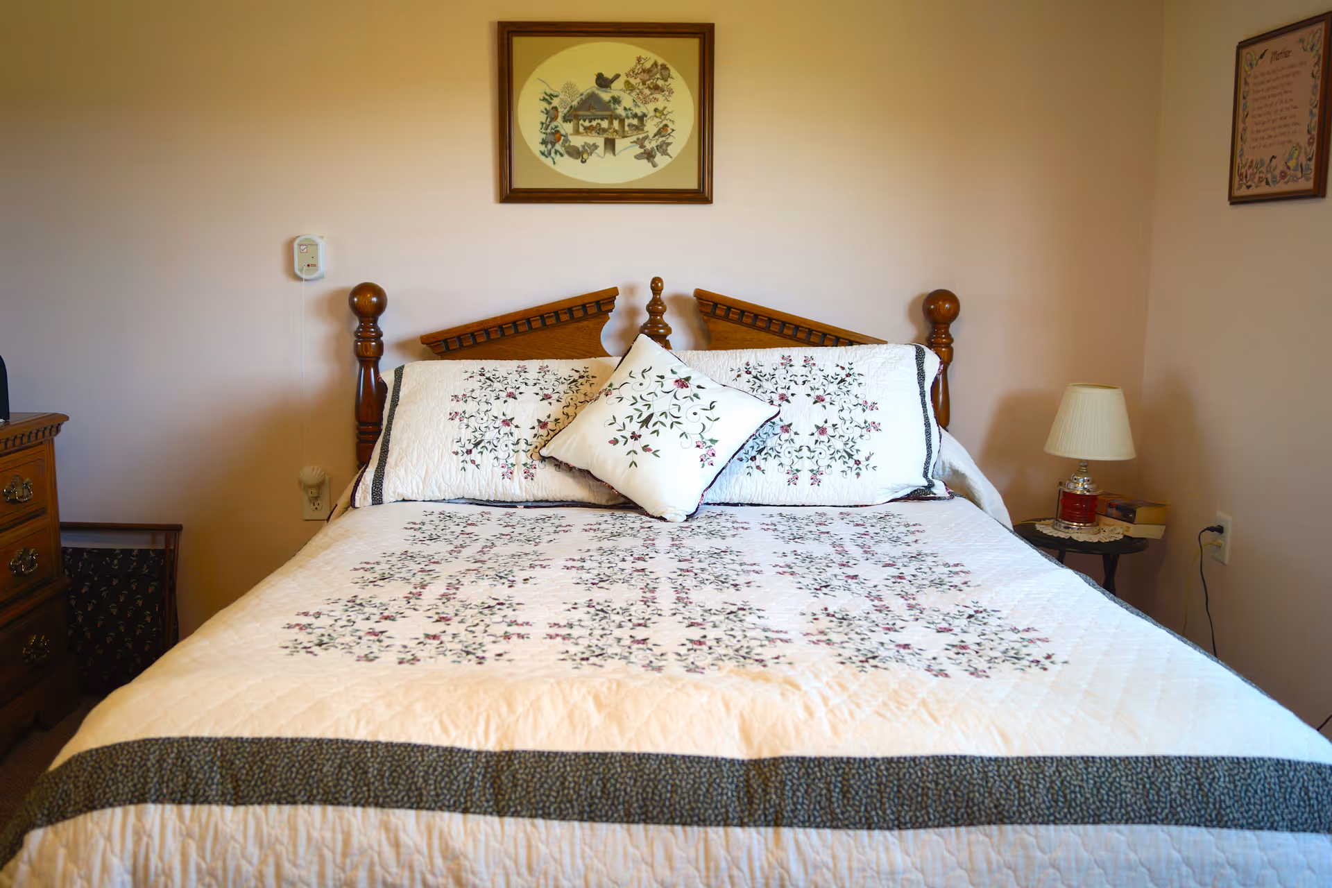A neatly made bed with a floral quilt and pillows in a softly lit bedroom with a wooden headboard and bedside tables.