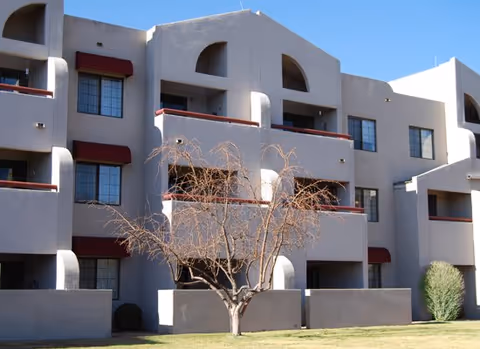 Beige three-story apartment building with recessed balconies, red awnings, and a small leafless tree on a grassy lawn.
