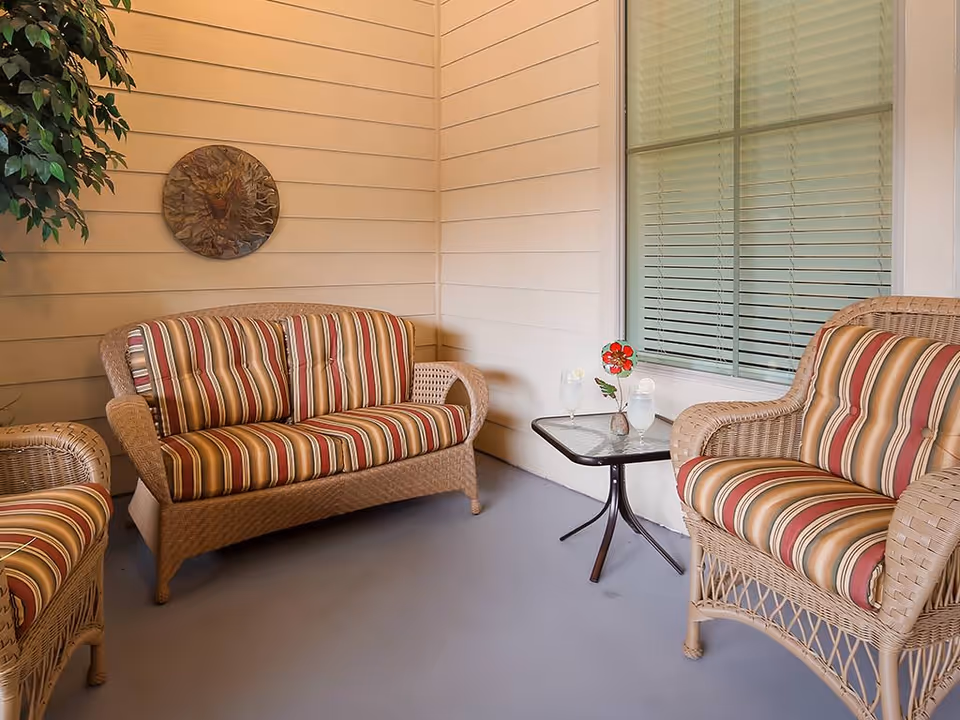 Wicker sofa and chairs with striped cushions on a covered porch with a small table holding drinks and a flower.
