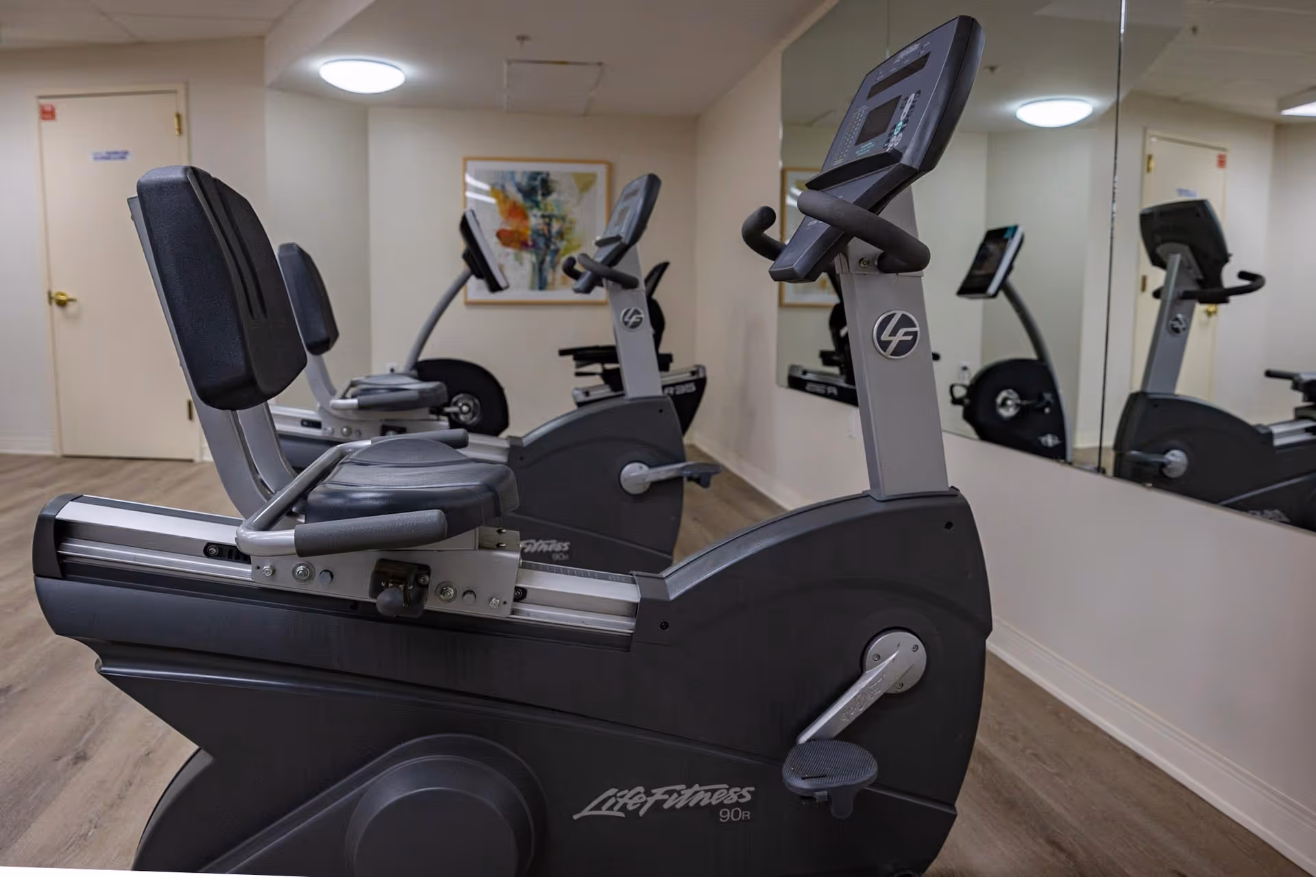 Two recumbent exercise bikes in a mirrored fitness room with wood flooring and neutral walls.