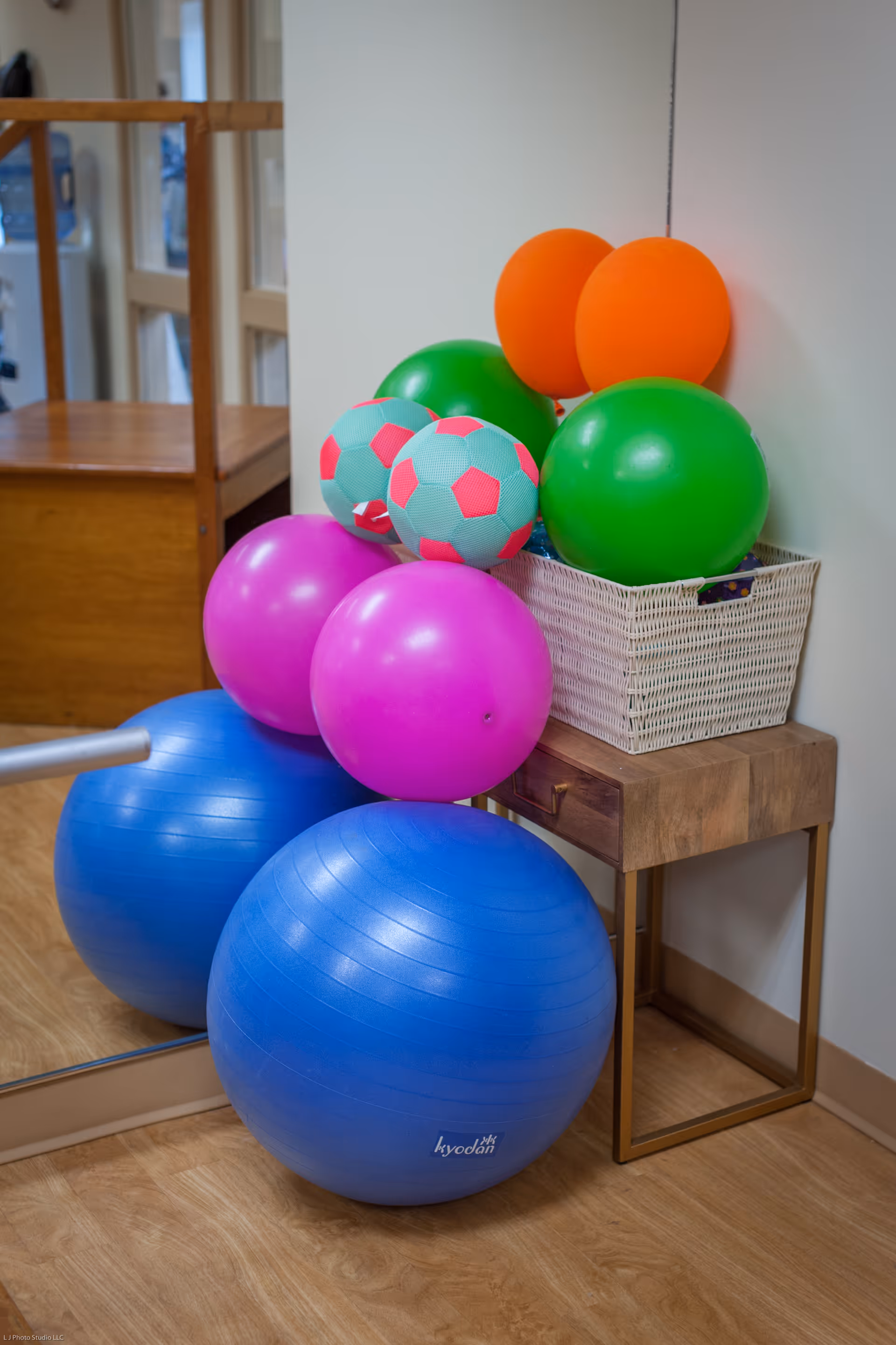 A corner of a room with a wooden floor and a mirror on the left wall. There is a small wooden table with a white wicker basket on top, holding several colorful balls including green, orange, and two blue and pink patterned balls. In front of the table are two large blue exercise balls and two pink balloons.