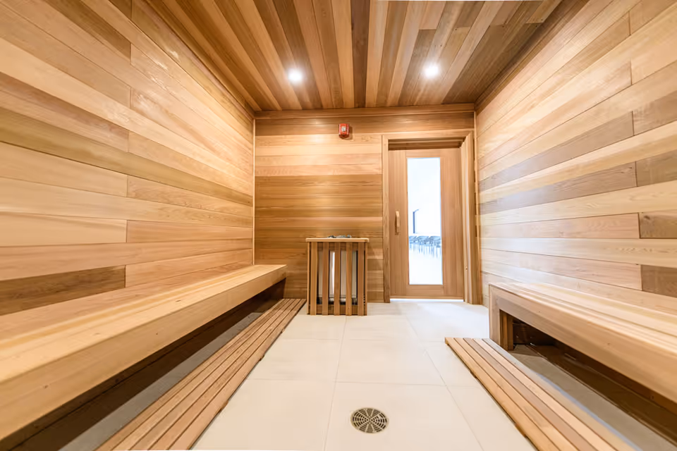 Interior view of a wooden sauna room with benches on both sides, a wooden heater guard in the center, and a glass door leading to a bright adjacent room.
