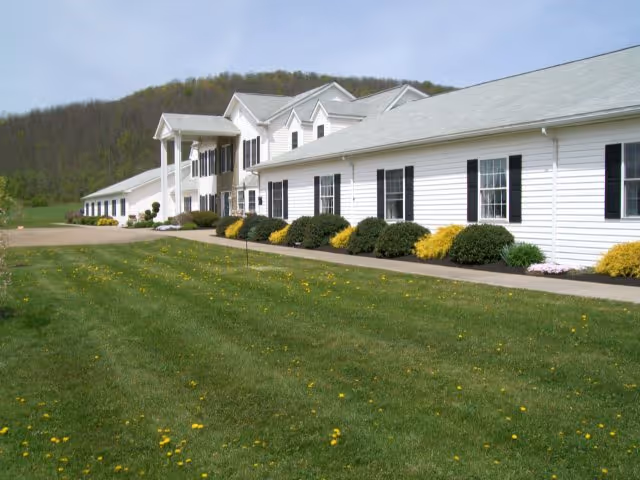 Exterior view of a long white building with black shutters and a gray roof, surrounded by green grass and bushes, with a hill in the background under a clear sky.