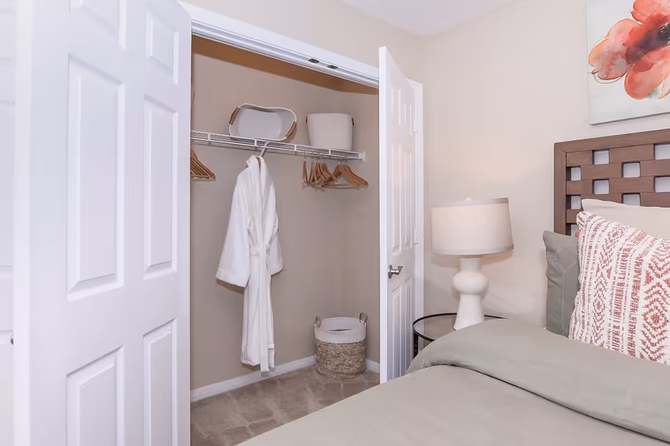 A bedroom corner showing an open closet with white double doors. Inside the closet, there are wooden hangers, a white bathrobe hanging, a white basket on the floor, and a tray and container on the top shelf. Next to the closet is a bedside table with a white lamp, and part of a bed with green bedding and patterned pillows is visible. A floral painting is partially visible on the wall above the bed.