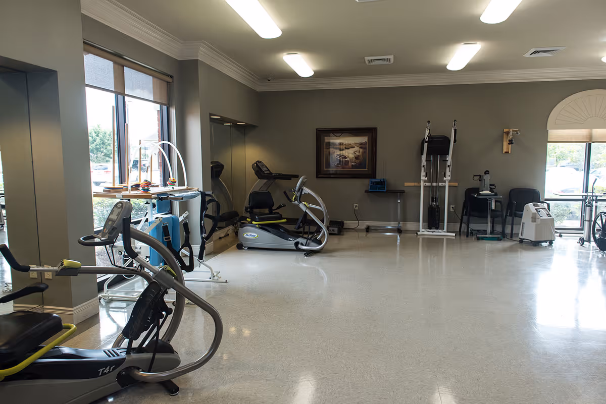 A spacious exercise room with various fitness equipment including stationary bikes, a treadmill, and other exercise machines. The room has large windows letting in natural light, gray walls, and a framed picture on the wall.
