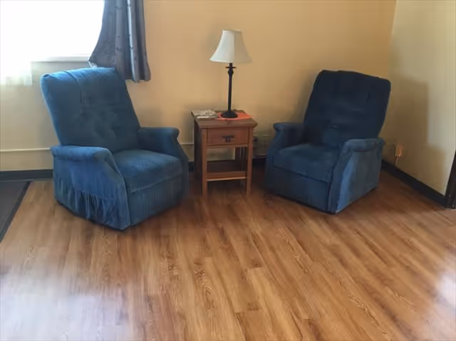 Two blue upholstered recliner chairs flanking a wooden side table with a lamp on wood-look flooring.