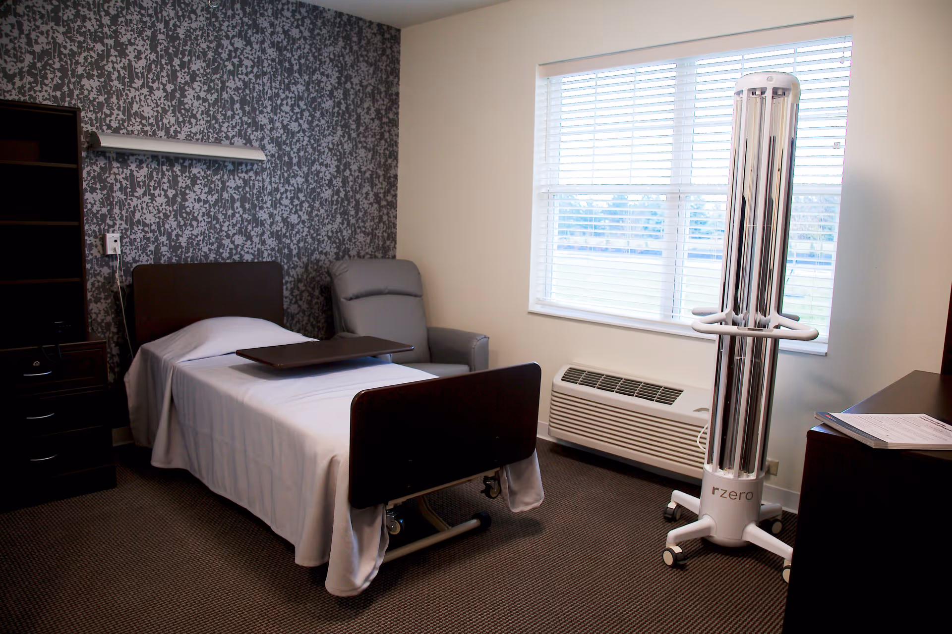 A small, clean bedroom in a healthcare facility with a single hospital bed covered with white sheets, a gray reclining chair beside the bed, a dark wooden nightstand and dresser, a window with blinds letting in natural light, and a tall white UV disinfection lamp labeled 'rzero' on wheels near the window.