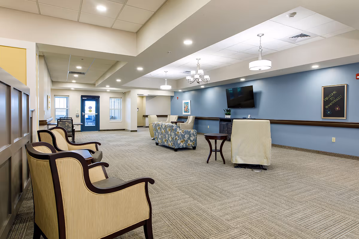 A spacious and well-lit common area in an assisted living facility with beige and floral upholstered chairs arranged around small tables. The walls are painted blue and beige, with a flat-screen TV mounted on the wall and a chalkboard sign that says 'Welcome to Homestead!'. The ceiling has recessed lighting and hanging light fixtures, and there are windows and a blue door at the far end of the room.