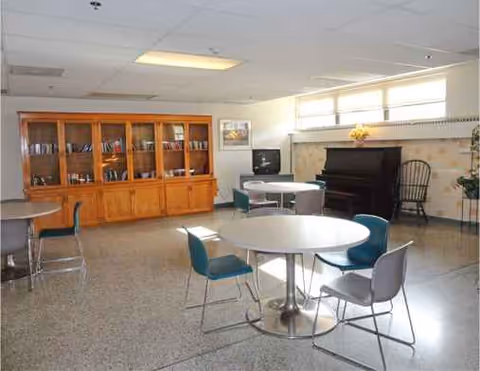 Common indoor lounge with round tables, chairs, a wooden cabinet, piano, and bright windows.