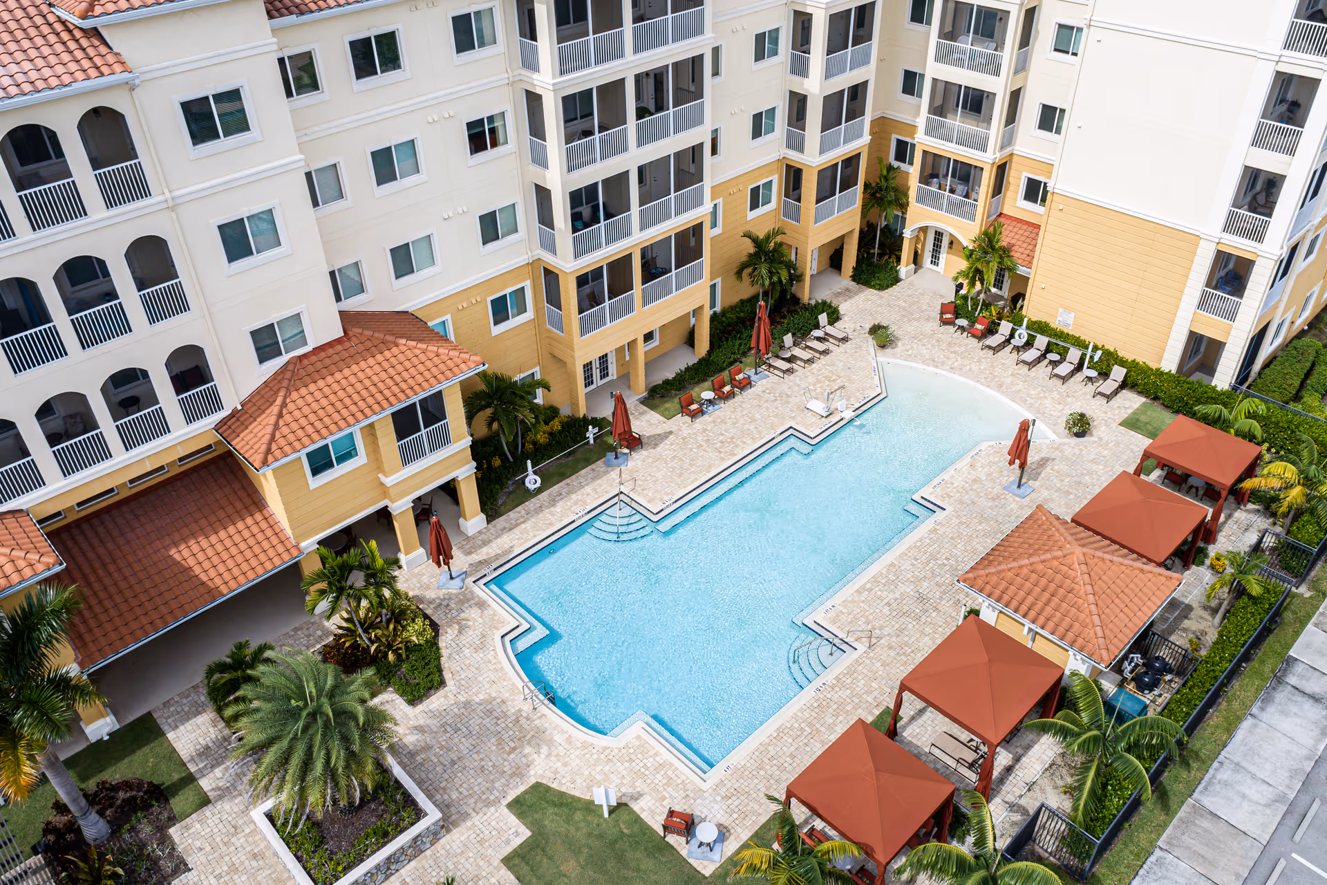 Aerial view of an outdoor swimming pool area surrounded by a multi-story residential building with balconies. The pool is rectangular with steps on two sides and is bordered by a tiled deck with lounge chairs, umbrellas, and cabanas. Palm trees and landscaped greenery are also visible around the pool area.