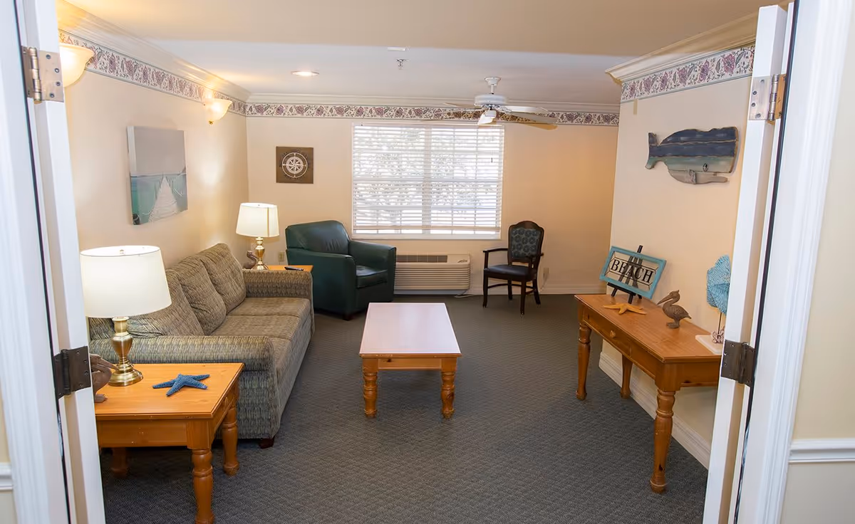 A cozy living room with a gray couch, a green armchair, and a wooden coffee table in the center. There is a wooden side table with a lamp and a blue starfish decoration on the left. On the right, a wooden console table holds a 'BEACH' sign, a pelican figurine, and other decorative items. The walls are light-colored with a floral border near the ceiling. A window with blinds lets in natural light, and a ceiling fan is mounted above.