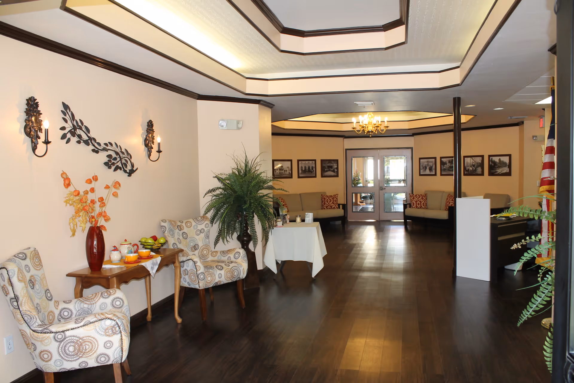 A spacious and well-lit interior lounge area with dark wood flooring and beige walls. The room features patterned armchairs, a small wooden table with a red vase holding orange flowers, and a tea set. Decorative wall sconces and metal wall art adorn the walls. In the background, there are beige sofas with patterned cushions, framed black and white photos on the walls, a chandelier, and double glass doors leading outside. There are also several green plants placed around the room.