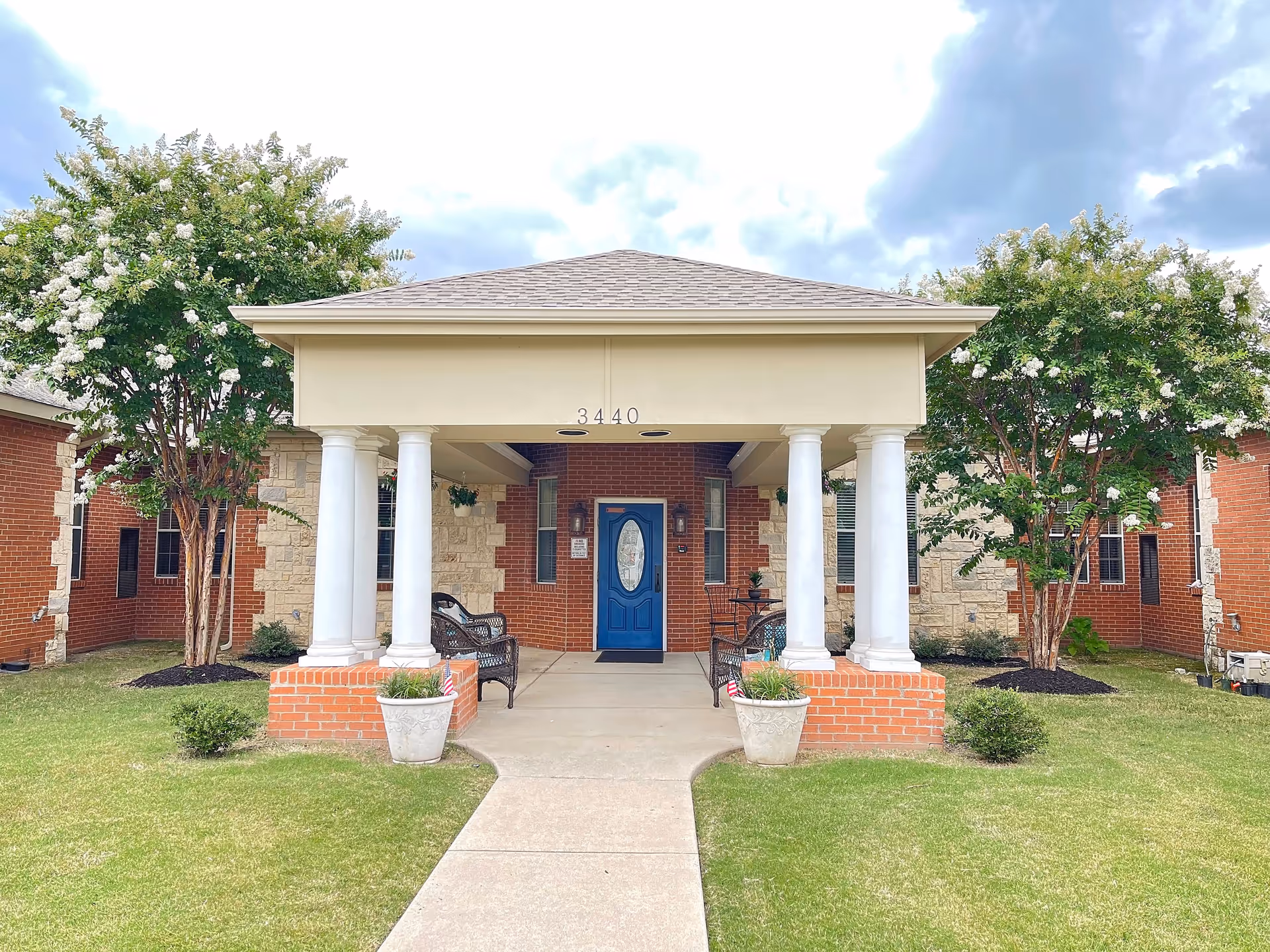 Front exterior view of a single-story brick and stone building with a covered entrance supported by four white columns. The entrance has a blue door with an oval glass window and is flanked by two windows. There are two potted plants on either side of the walkway leading to the door, and two flowering trees on each side of the building. The building number 3440 is displayed above the entrance.