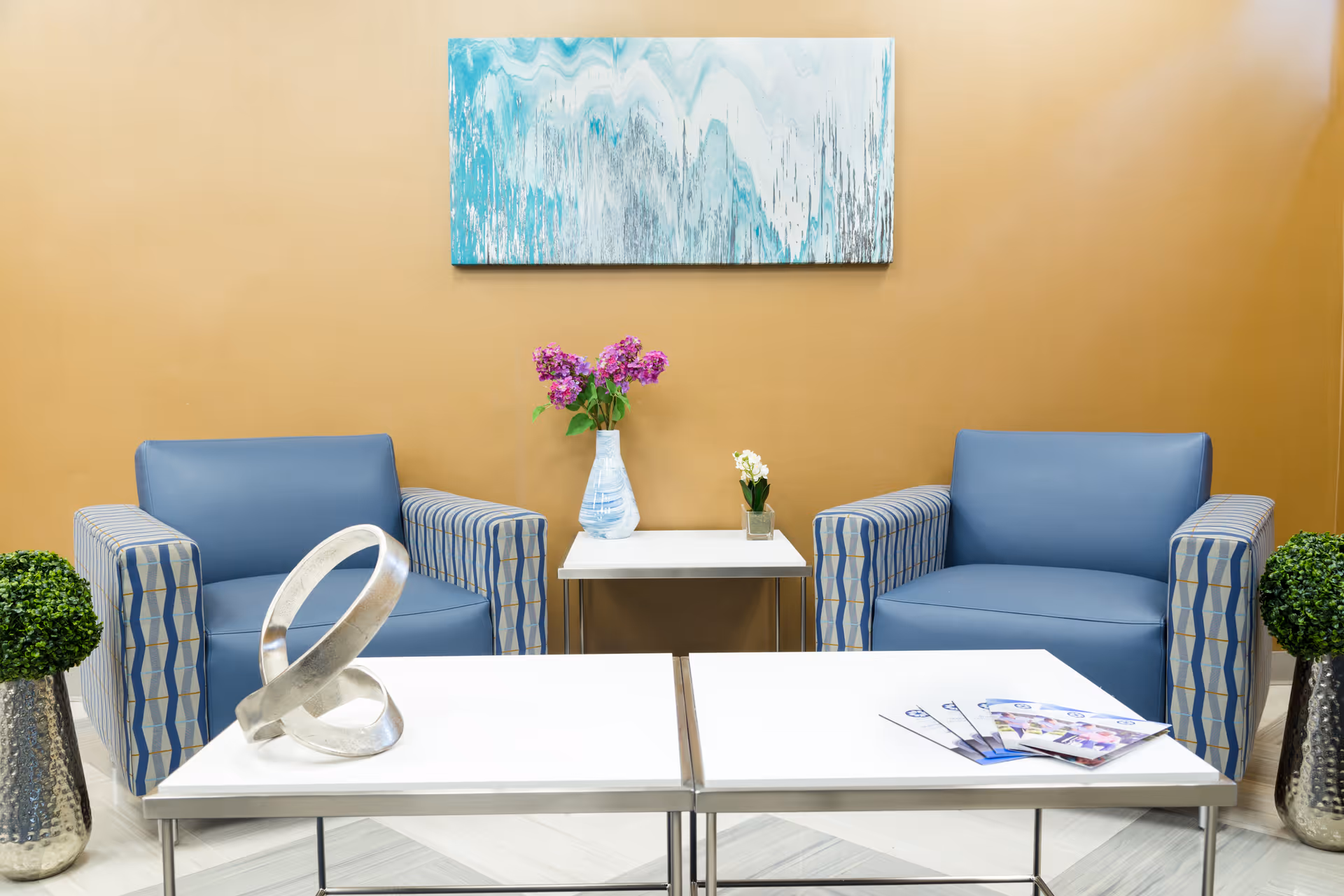 Two blue armchairs face a white coffee table in a clean waiting area with vases of flowers and artwork on the wall.