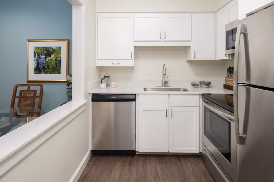 A modern kitchen with white cabinets, a stainless steel dishwasher, oven, microwave, and refrigerator. The kitchen has a single sink with a faucet and a white countertop. To the left, there is a partial view of a dining area with a glass table, wooden chairs, a blue accent wall, and a framed picture of grapes.