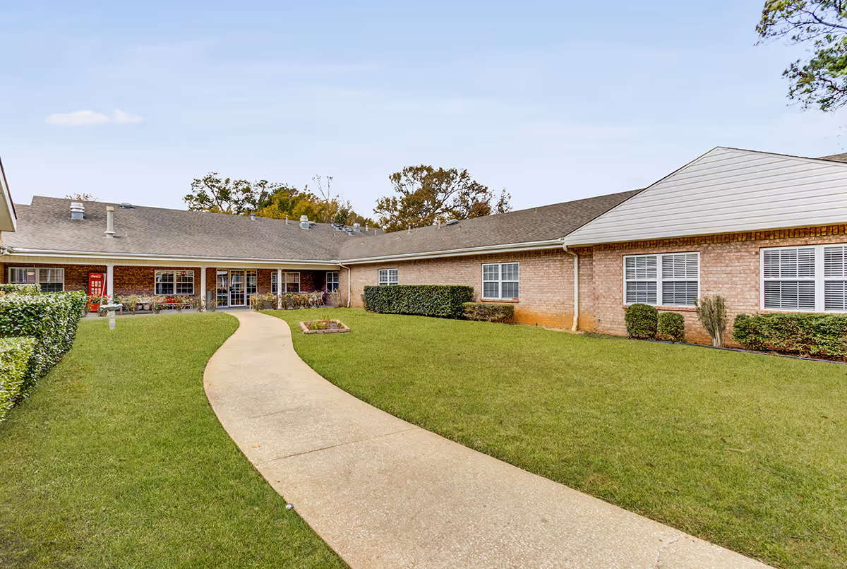 Brick senior living facility building with a curved concrete walkway through a grassy courtyard leading to the entrance.