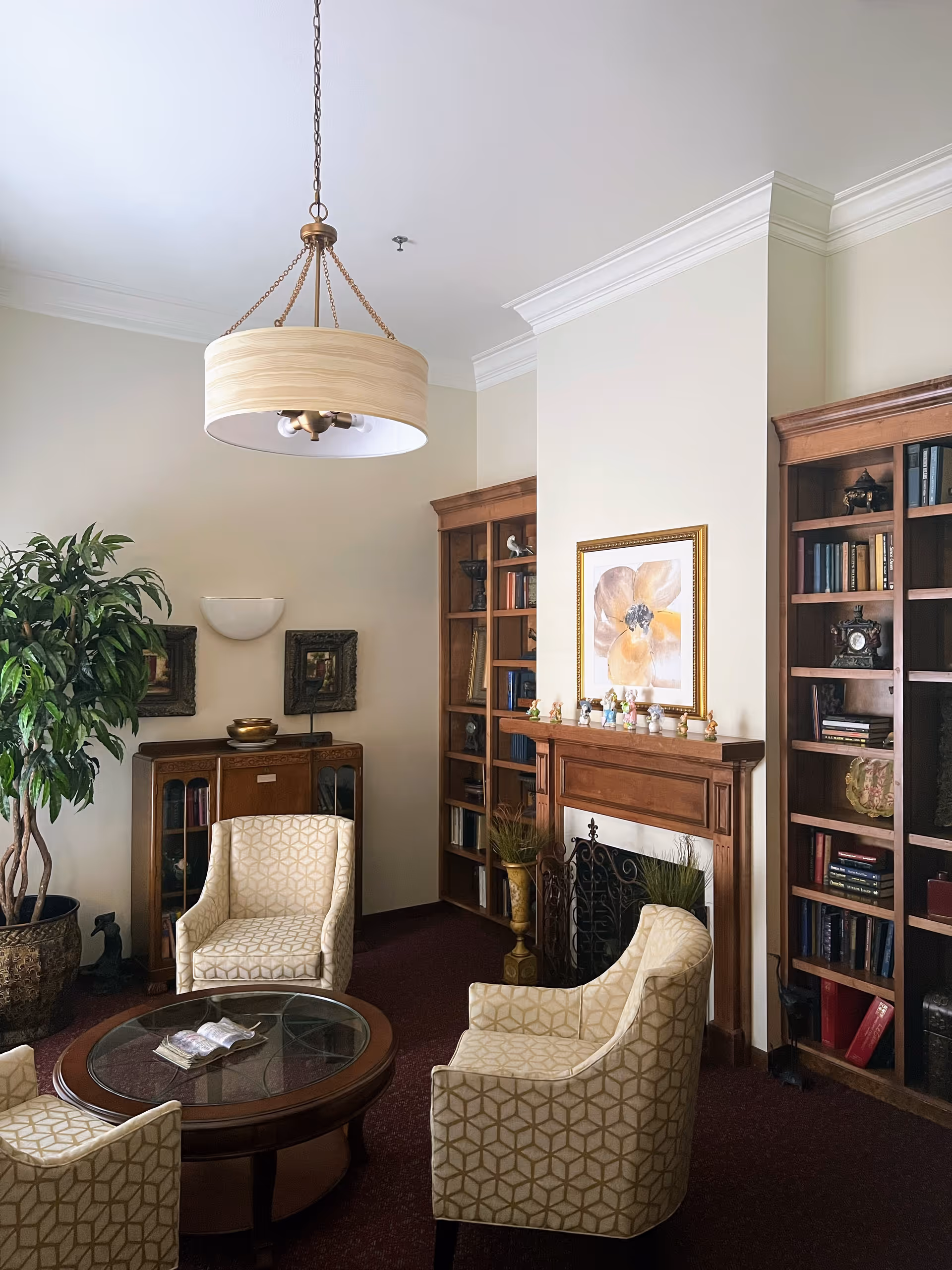 A cozy sitting area in a senior living facility with four patterned armchairs arranged around a round glass-top wooden coffee table. The room features a wooden fireplace mantel decorated with small figurines and a large framed flower painting above it. There are built-in wooden bookshelves filled with books and decorative items on either side of the fireplace. A large potted plant is placed in the corner, and a modern chandelier hangs from the ceiling.