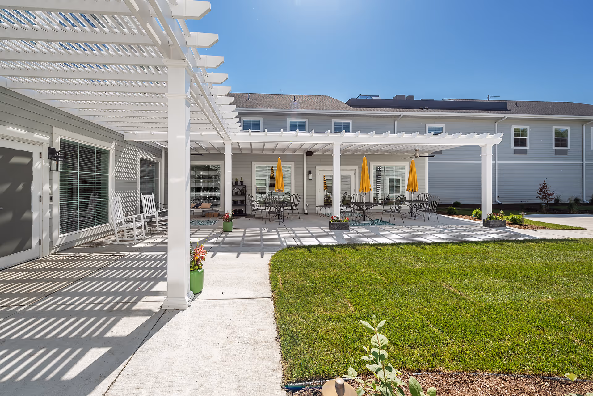 Outdoor patio area at Provision Living at East Lansing featuring a white pergola casting shadows on the concrete floor, several round tables with yellow umbrellas, black metal chairs, white rocking chairs, potted plants, and a well-maintained green lawn under a clear blue sky.
