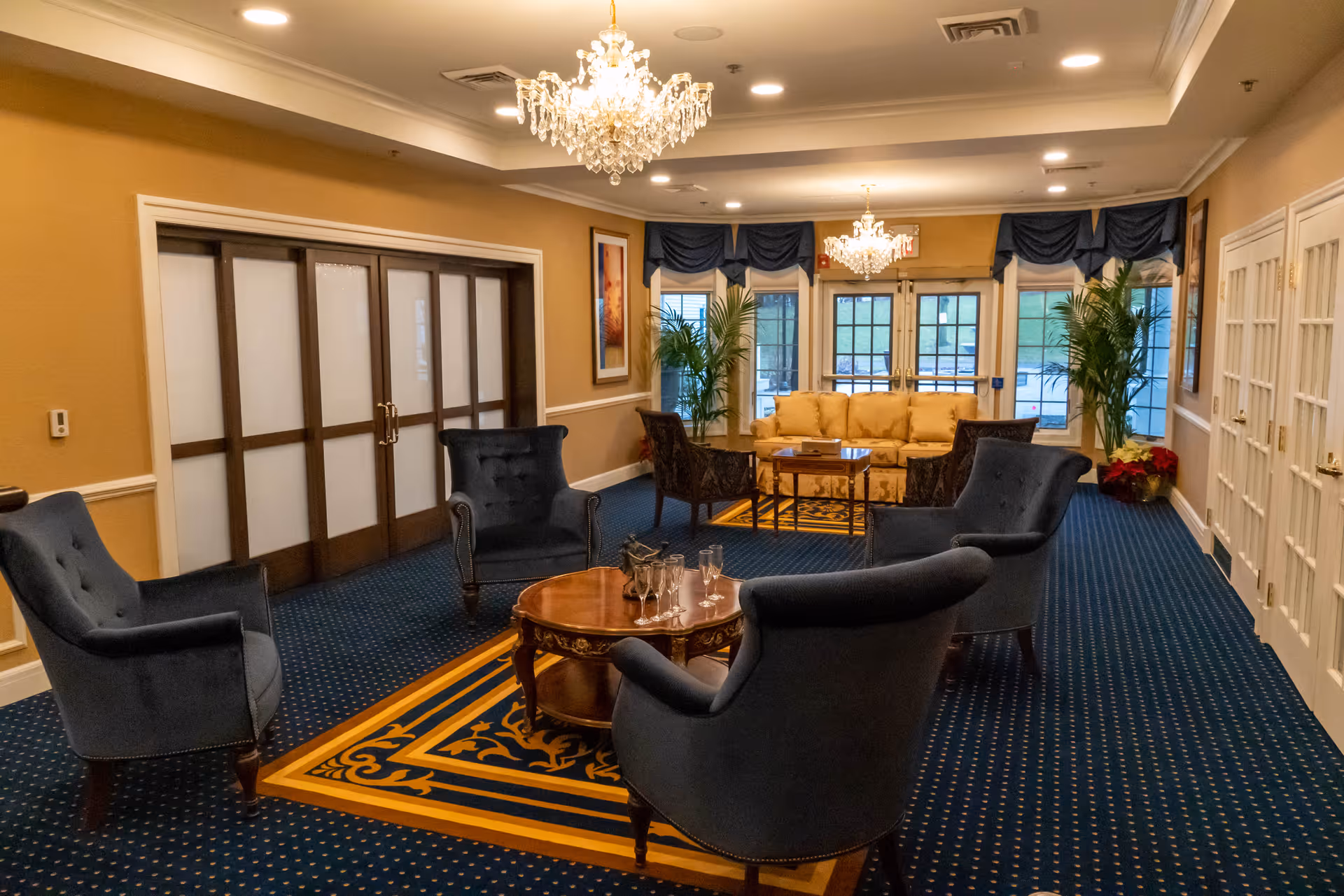 A cozy and elegant lounge area with four blue upholstered armchairs arranged around a wooden coffee table with glasses and a pitcher. In the background, there is a beige sofa with a glass coffee table in front of it, flanked by two large potted plants. The room has blue carpet with a decorative yellow and blue rug, beige walls, two chandeliers, and large windows with blue valances.