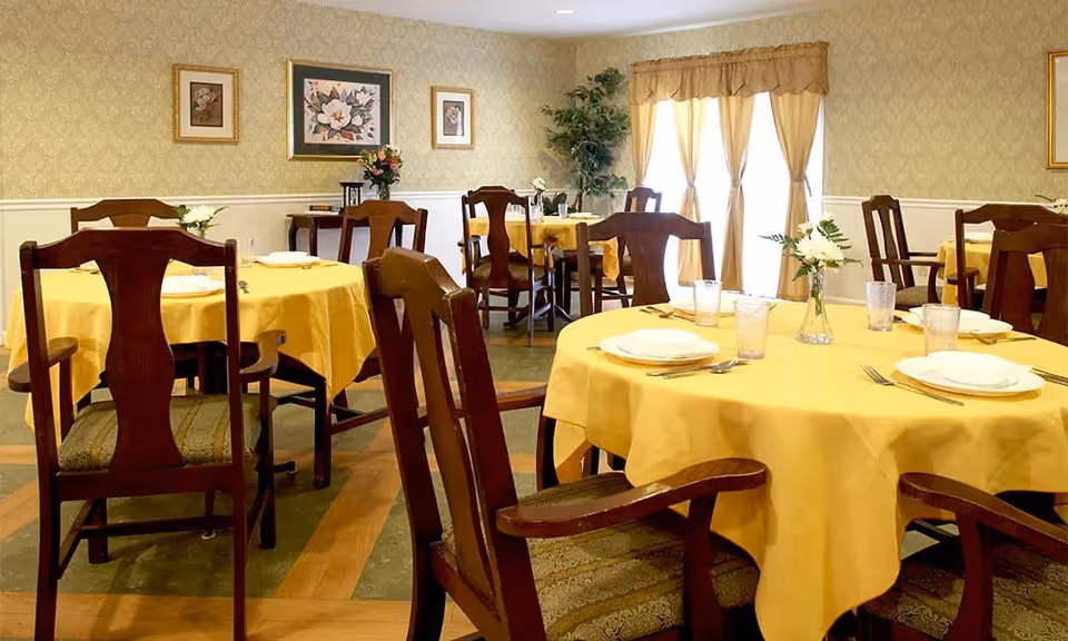 A dining room with round tables covered in yellow tablecloths, each set with plates, glasses, and silverware. Wooden chairs with cushioned seats surround the tables. The room has patterned wallpaper, framed floral artwork on the walls, and a window with beige curtains letting in natural light. A small plant is placed in the corner.