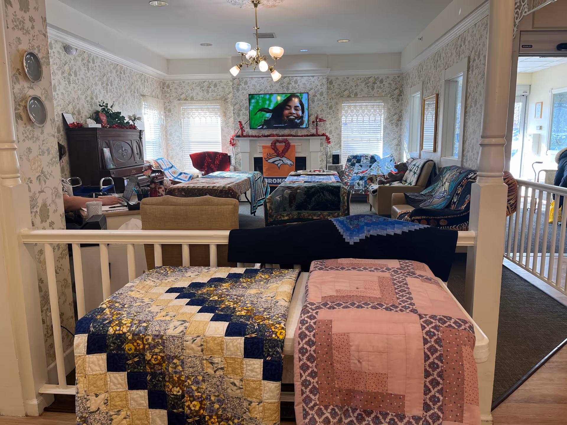 A cozy senior living common room with quilts draped over a railing in the foreground, seating arranged around a fireplace with a TV mounted above, and floral wallpapered walls.