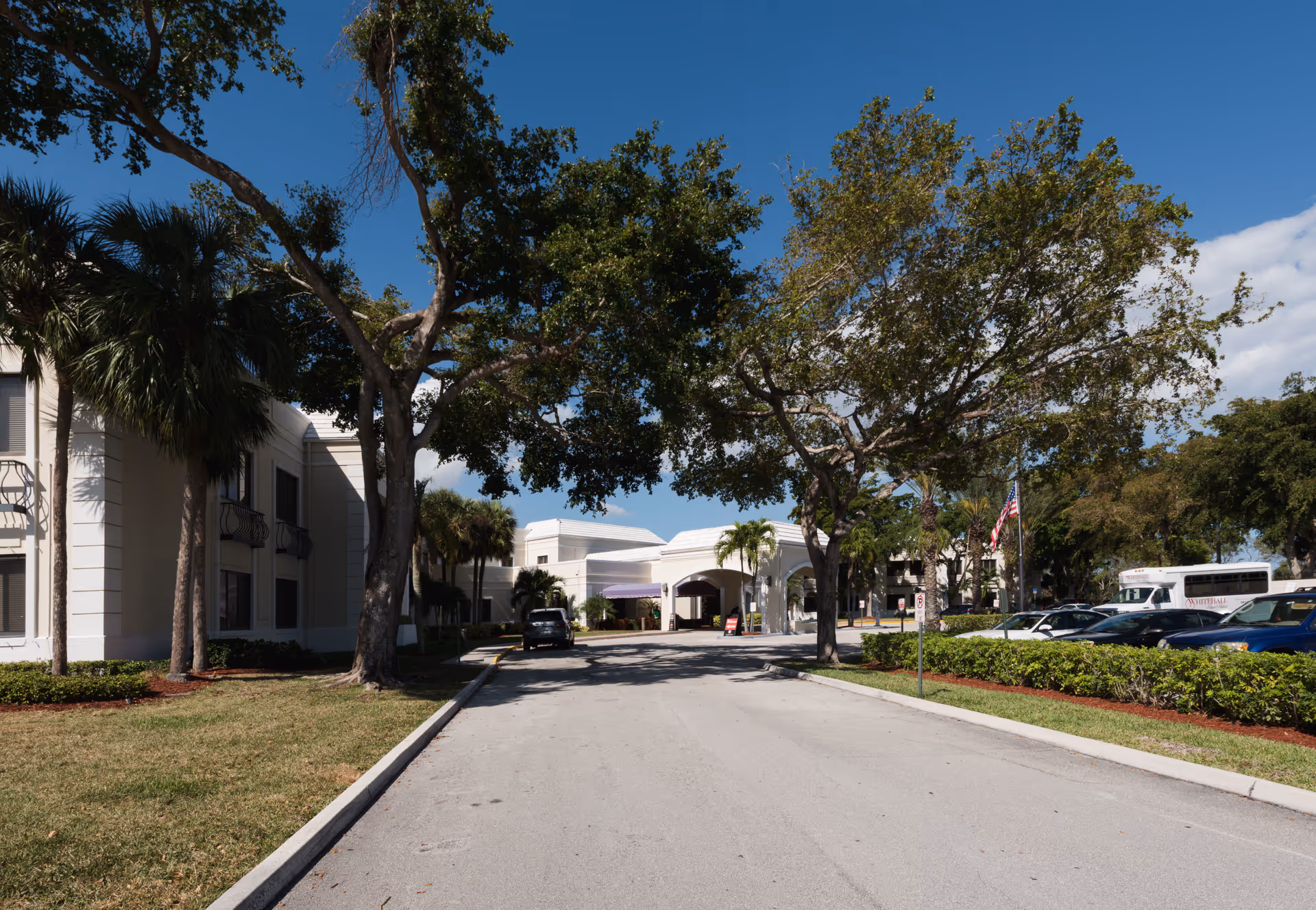 Driveway leading to the front entrance of a low-rise care facility flanked by trees, parked cars, and a shuttle bus under a blue sky.