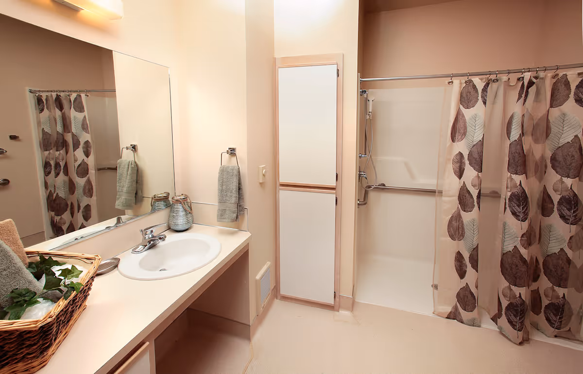Bright bathroom with a sink and vanity on the left, a tall cabinet in the center, and a shower with a leaf-patterned curtain on the right.