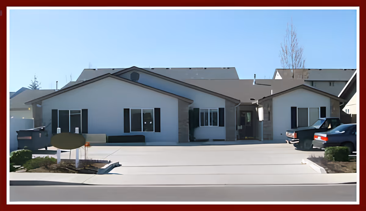 Exterior view of a single-story building with white walls and dark window shutters, a driveway in front with parked cars on the right side, and a clear blue sky above.