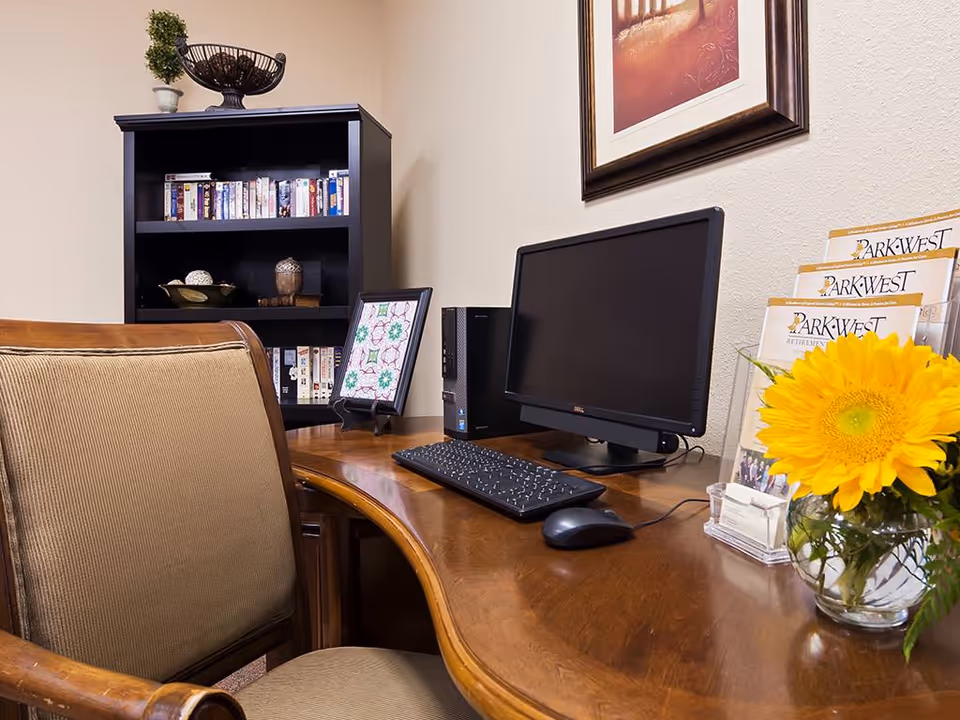 A cozy office corner with a wooden desk holding a computer monitor, keyboard, and mouse. A vase with a bright yellow sunflower and brochures labeled 'Park West' are on the desk. Behind the desk is a black bookshelf filled with books and decorative items. A framed picture hangs on the wall above the desk, and a cushioned wooden chair is positioned in front of the desk.