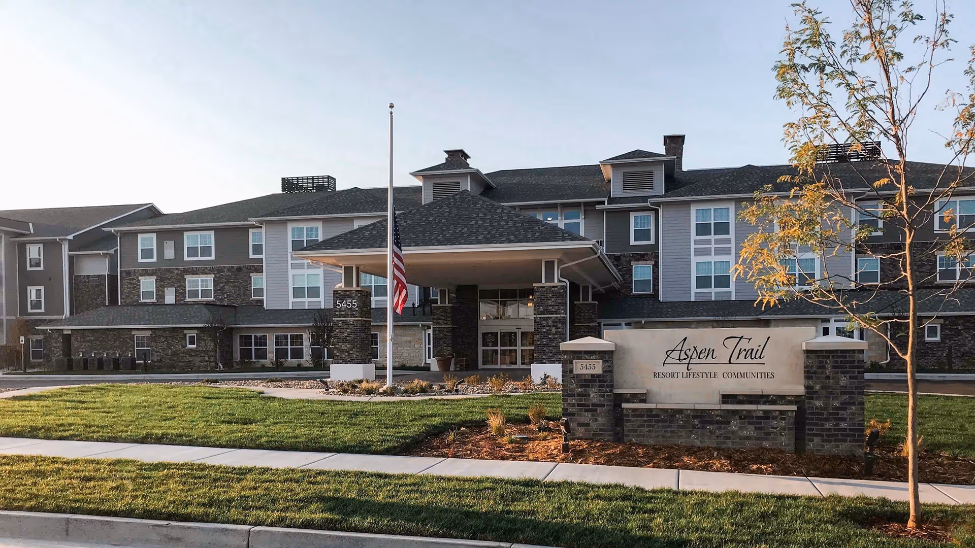 Front exterior of the Aspen Trail Retirement Resort building with an entrance canopy, flagpole, and a sign on a landscaped lawn.