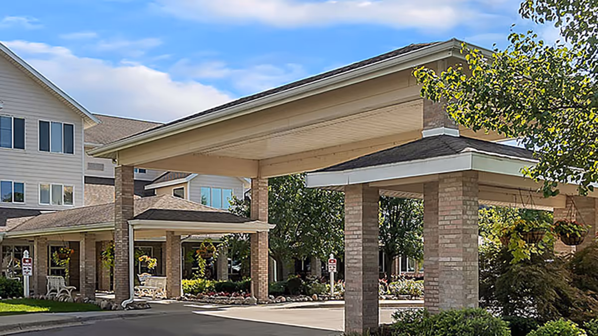 Exterior view of Holiday Genesee Gardens senior living facility showing a covered entrance with brick pillars, hanging flower baskets, and a multi-story building in the background under a partly cloudy sky.