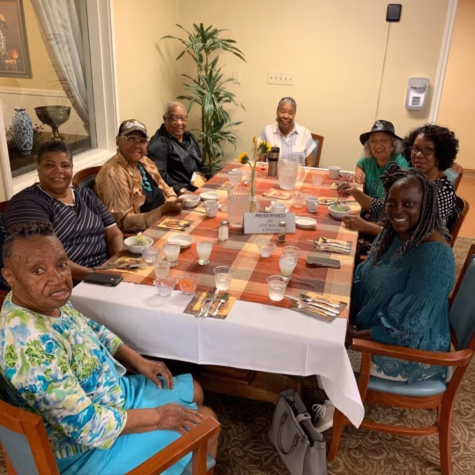 A group of eight elderly women sitting around a rectangular dining table with a checkered tablecloth, enjoying a meal together in a well-lit room with a potted plant in the corner. The table is set with plates, cups, utensils, and a reserved sign in the center.