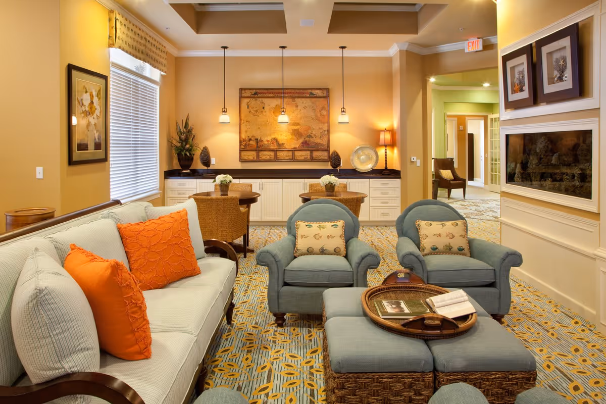 A cozy living room area with a light blue sofa adorned with orange and light blue pillows, two matching armchairs with patterned cushions, and a woven ottoman with a tray holding books. The room features warm yellow walls, a patterned carpet, a wall-mounted electric fireplace, and a sideboard with decorative items and hanging pendant lights. In the background, there is a small dining area with wicker chairs and a framed map on the wall.