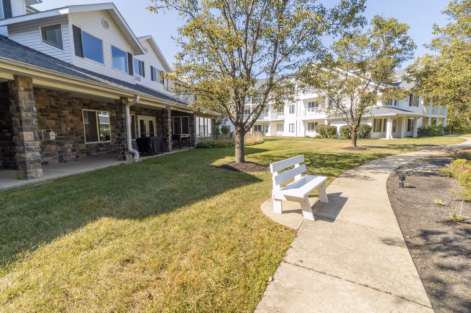 Outdoor view of a senior living facility with a paved walkway, a white bench, green grass, trees, and multi-story buildings with balconies and stone facades under a clear sky.