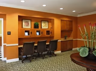 Interior business center with three computer stations in a wooden built-in desk, three rolling chairs, patterned carpet, and a round table with plants.