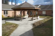 Outdoor courtyard area of a senior living facility with a wooden gazebo at the center, surrounded by a concrete walkway and grass. The building with beige walls and multiple windows encloses the courtyard.