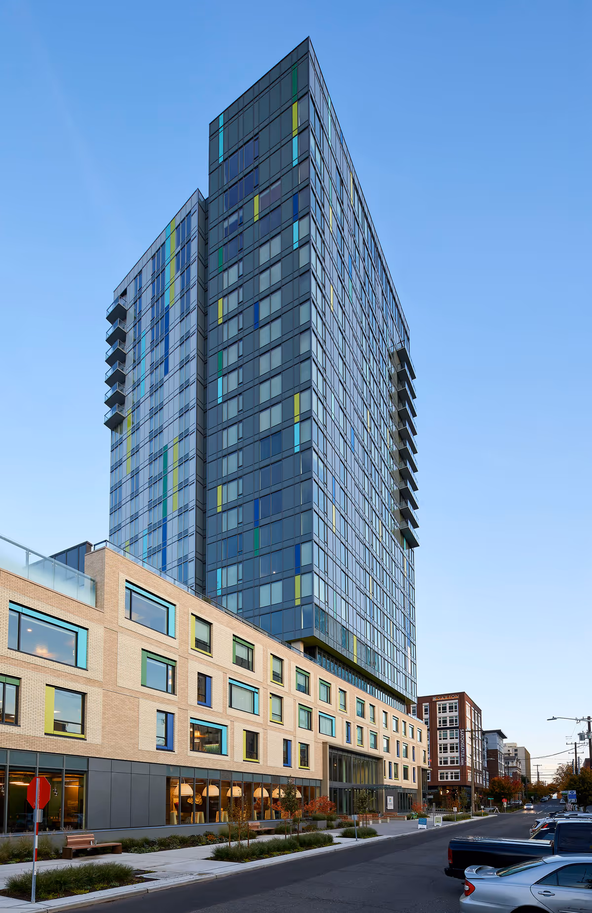 Tall modern senior living facility building with a glass and brick exterior, featuring colorful vertical accents on the glass windows, situated along a street with parked cars and a clear blue sky.