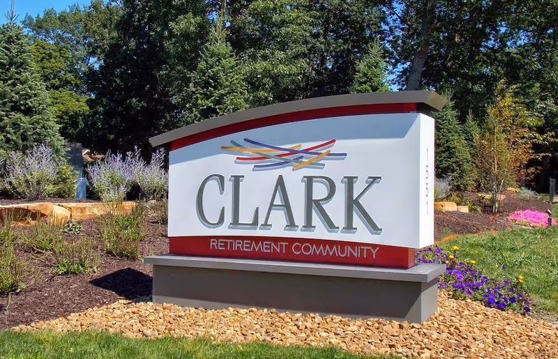 Entrance sign reading 'CLARK Retirement Community' amid landscaped beds, flowers, and trees.